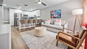 Una sala de estar moderna con sofá, una mesa de centro y sillones, una cocina al fondo y un ventilador de techo.