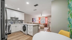 Una cocina moderna con armarios blancos, lavadora integrada y zona de comedor con mesa redonda y sillas amarillas.