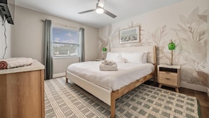 Un dormitorio con cama, mesitas de noche, ventilador de techo, ventana con cortinas y pared con diseño.
