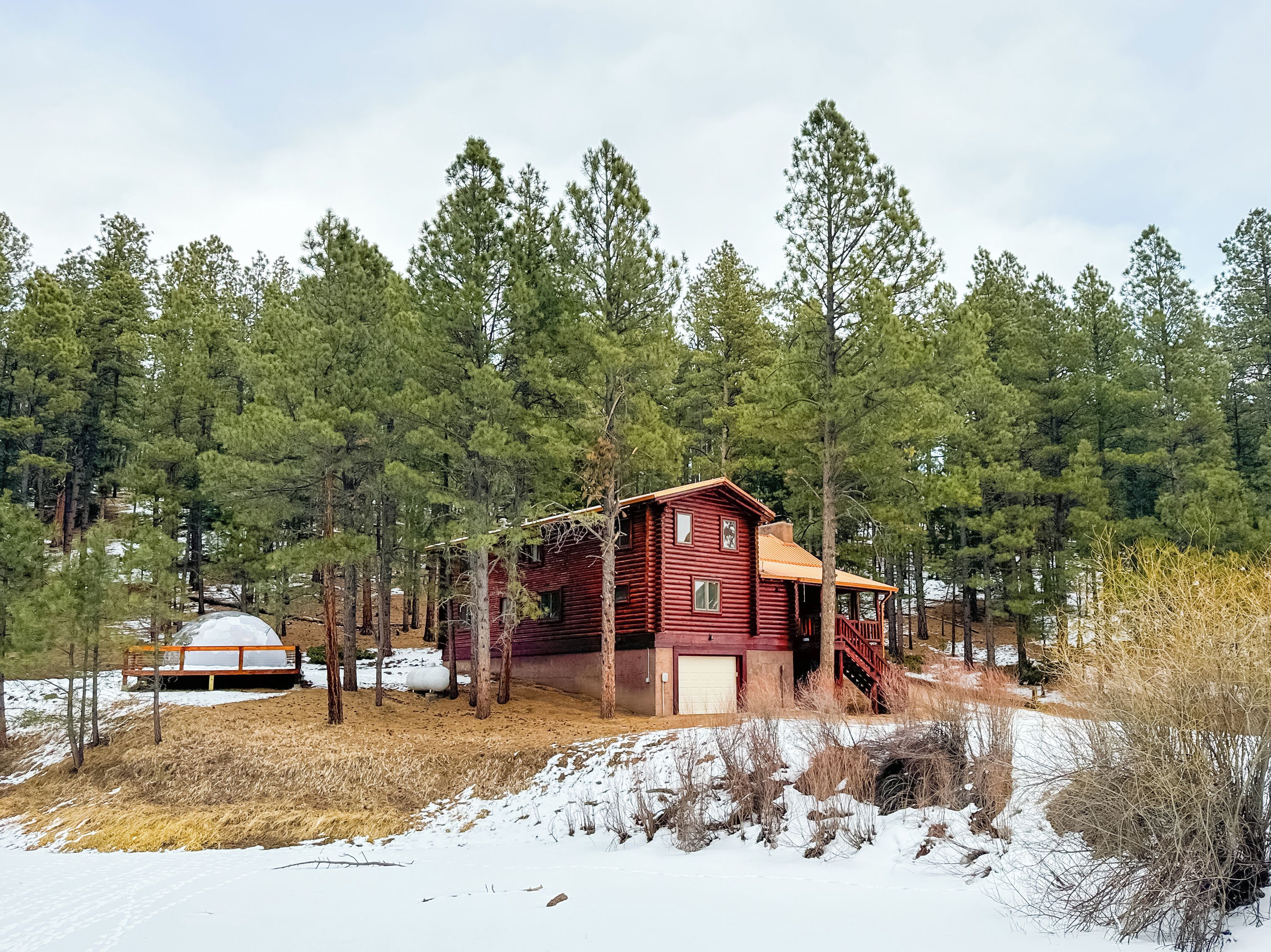 Una cabaña de troncos con un porche y terraza cubierta, rodeada de un bosque y nieve.