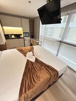Quarto de hotel com cama, televisão presa na parede, cozinha compacta e uma janela grande.