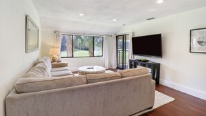 A modern living room with a beige sectional sofa, a round coffee table, and a flat-screen TV mounted on the wall. - Saddlebrook Golf Course Condo Voila Rent (Wesley Chapel)