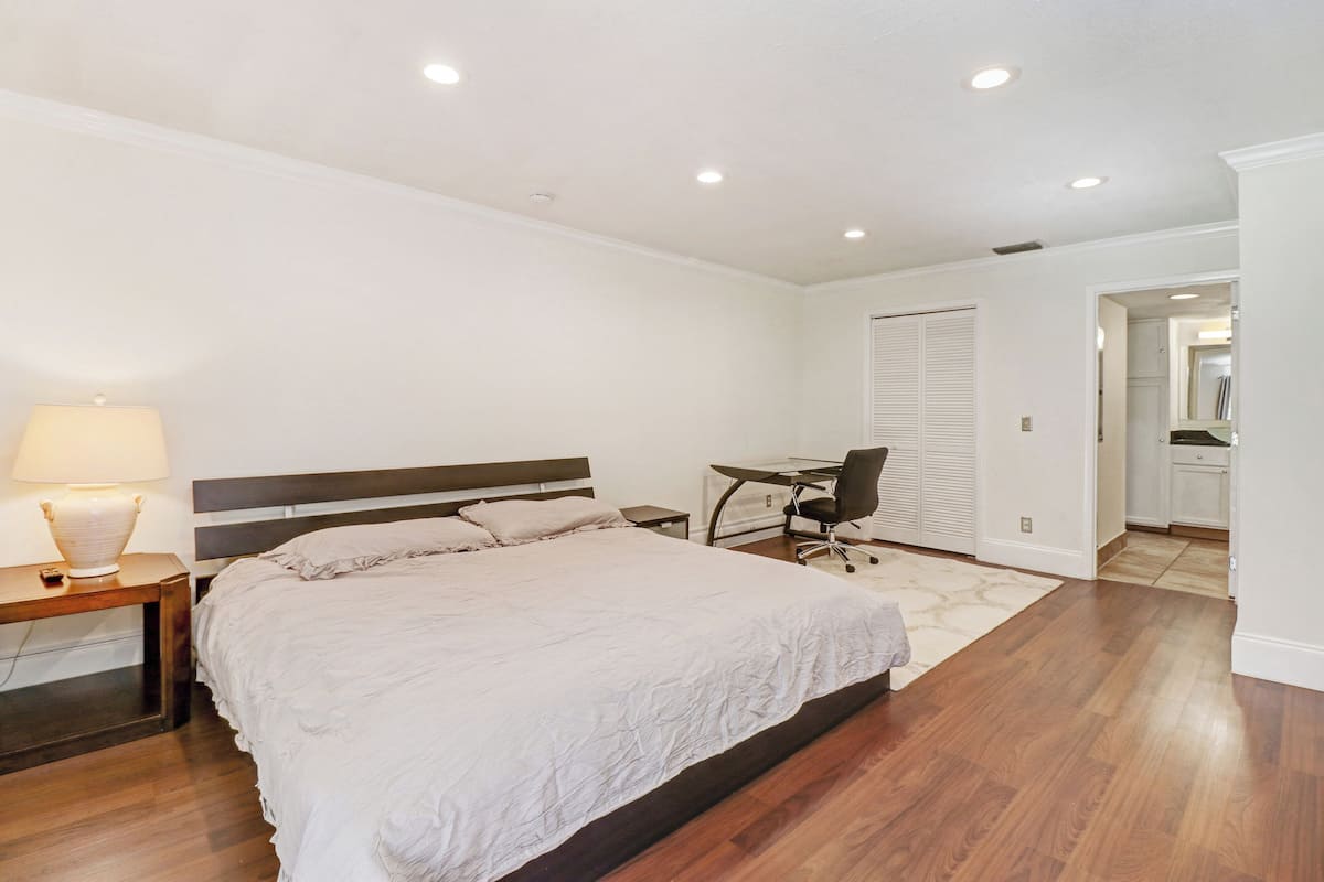 A bedroom with a large bed, a desk, and a chair. There is a nightstand with a lamp.