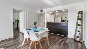 A modern dining area with a white table, teal plates, and white chairs. There is a wooden floor and a kitchen in the background. - Saddlebrook Golf Course Condo Voila Rent (Wesley Chapel)