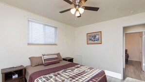 Ein Schlafzimmer mit einem Bett, einem Deckenventilator, einem Fenster mit Jalousien, einem gerahmten Bild an der Wand und einer Tür zu einem anderen Raum.