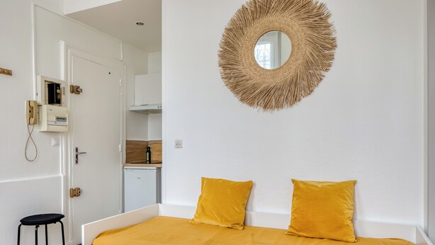 A bedroom with a bed, two yellow pillows, a white dresser, a small stool, and a wall-mounted mirror. - Urszula, charmant studio proche du Vieux Marché (Rouen)