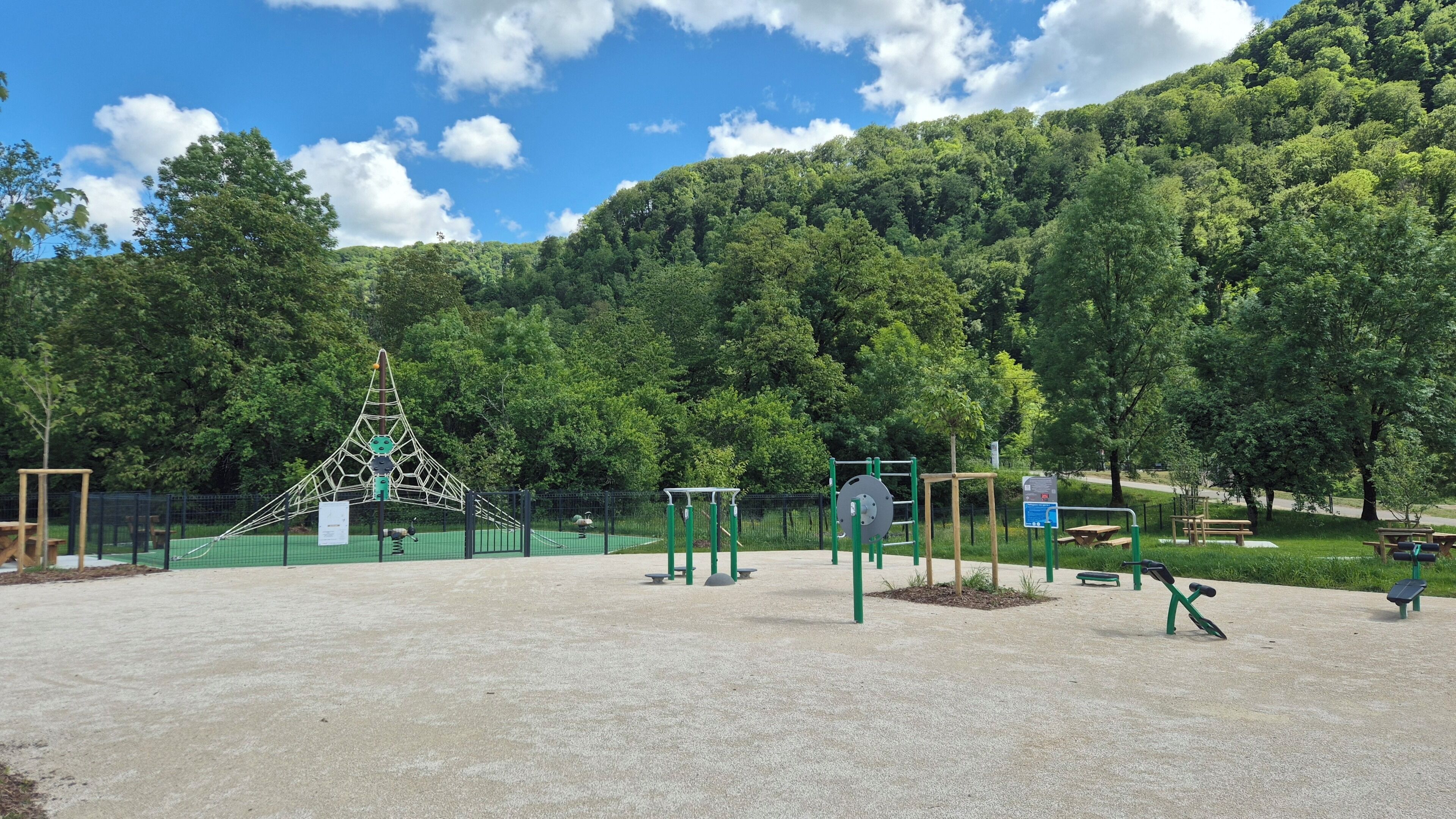 A playground with climbing structures, exercise equipment, and picnic tables in a park with a forested hill in the background.