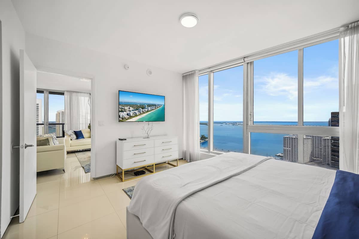 A modern hotel room with a large bed, a white dresser, a flat-screen TV mounted on the wall, and a large window offering a view of the sea and cityscape.