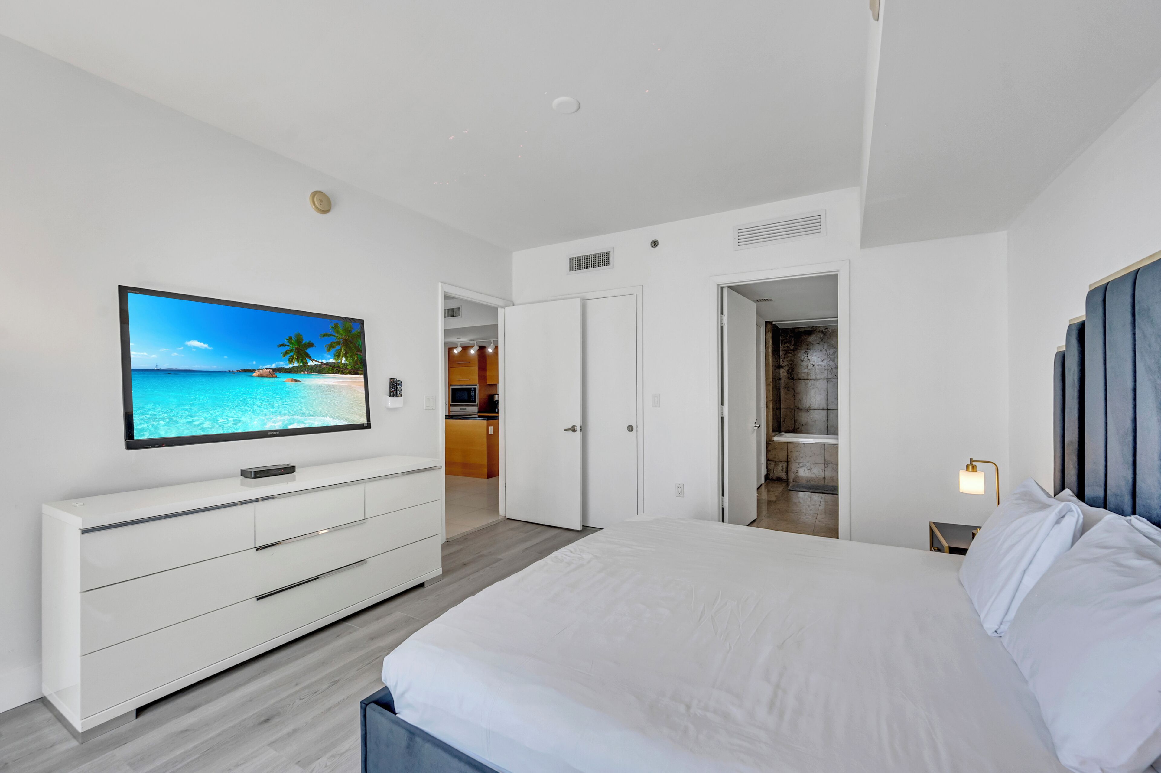 A modern hotel room with a large bed, a flat-screen TV on a white dresser, a walk-in closet, and a bathroom with a shower and bathtub.