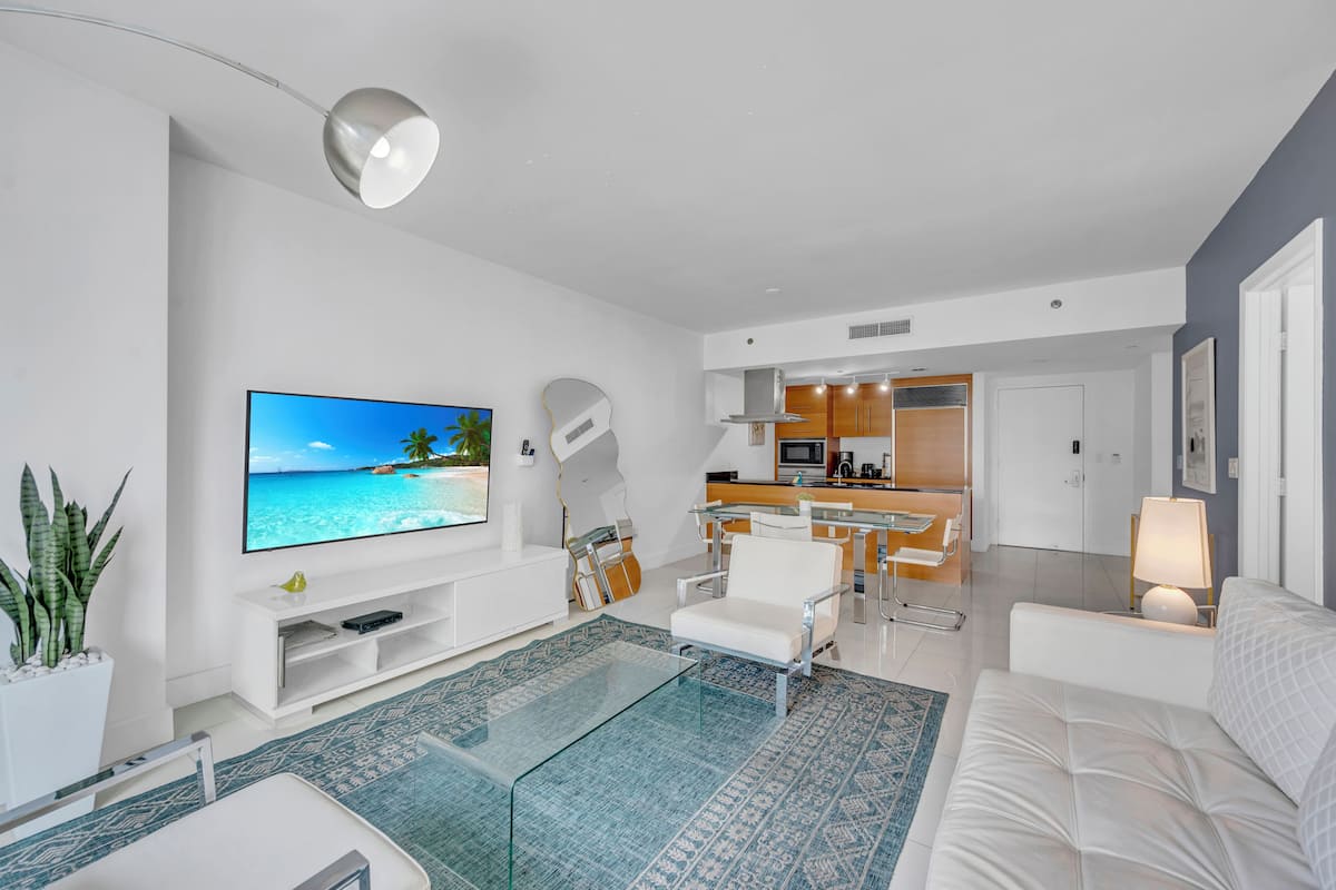 A modern living room with a flat-screen TV, a glass coffee table, a white sofa, and a kitchen area in the background.
