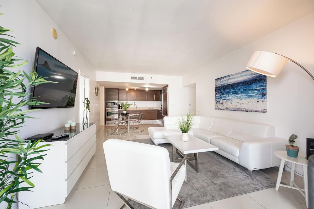A modern living room with a white sofa, a coffee table, and a TV. There is a kitchen in the background with a dining area.