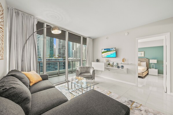 A modern living room with a grey sofa, a glass coffee table, and a view of high-rise buildings through large windows.