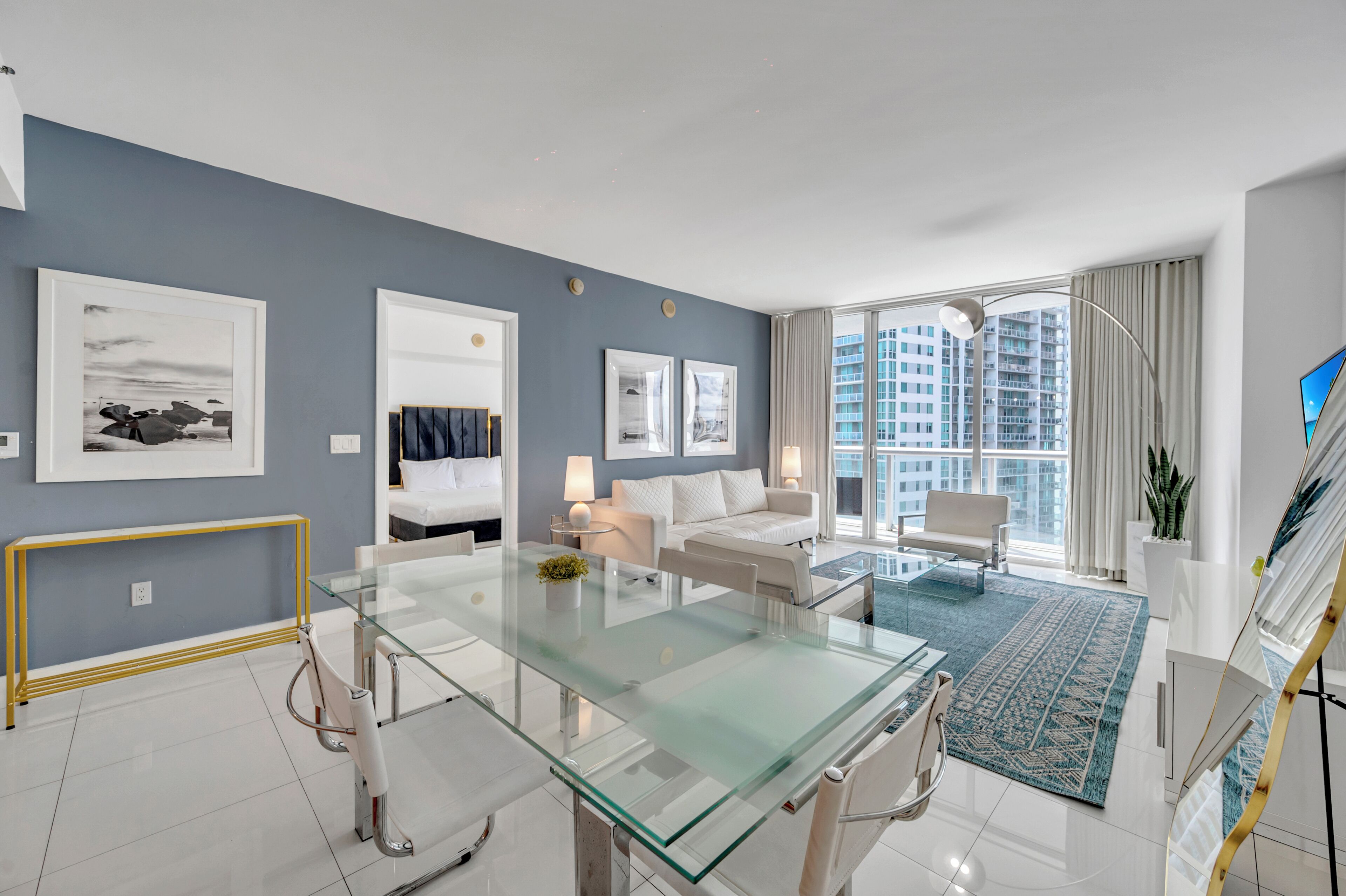A modern living room with a glass dining table, white sofas, and a large window with a city view.