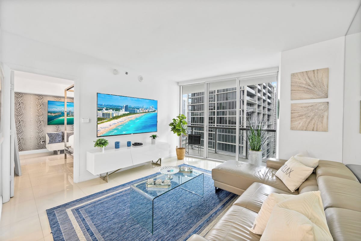 A modern living room with a large flat-screen TV, a glass coffee table, a beige sofa, and a large window with a city view.