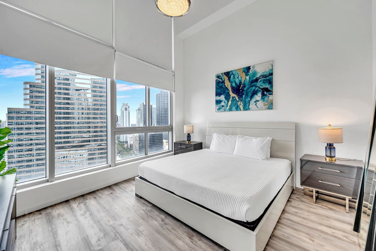 A modern bedroom with a large bed, a nightstand, a bedside table, a floor lamp, and a city view through large windows.