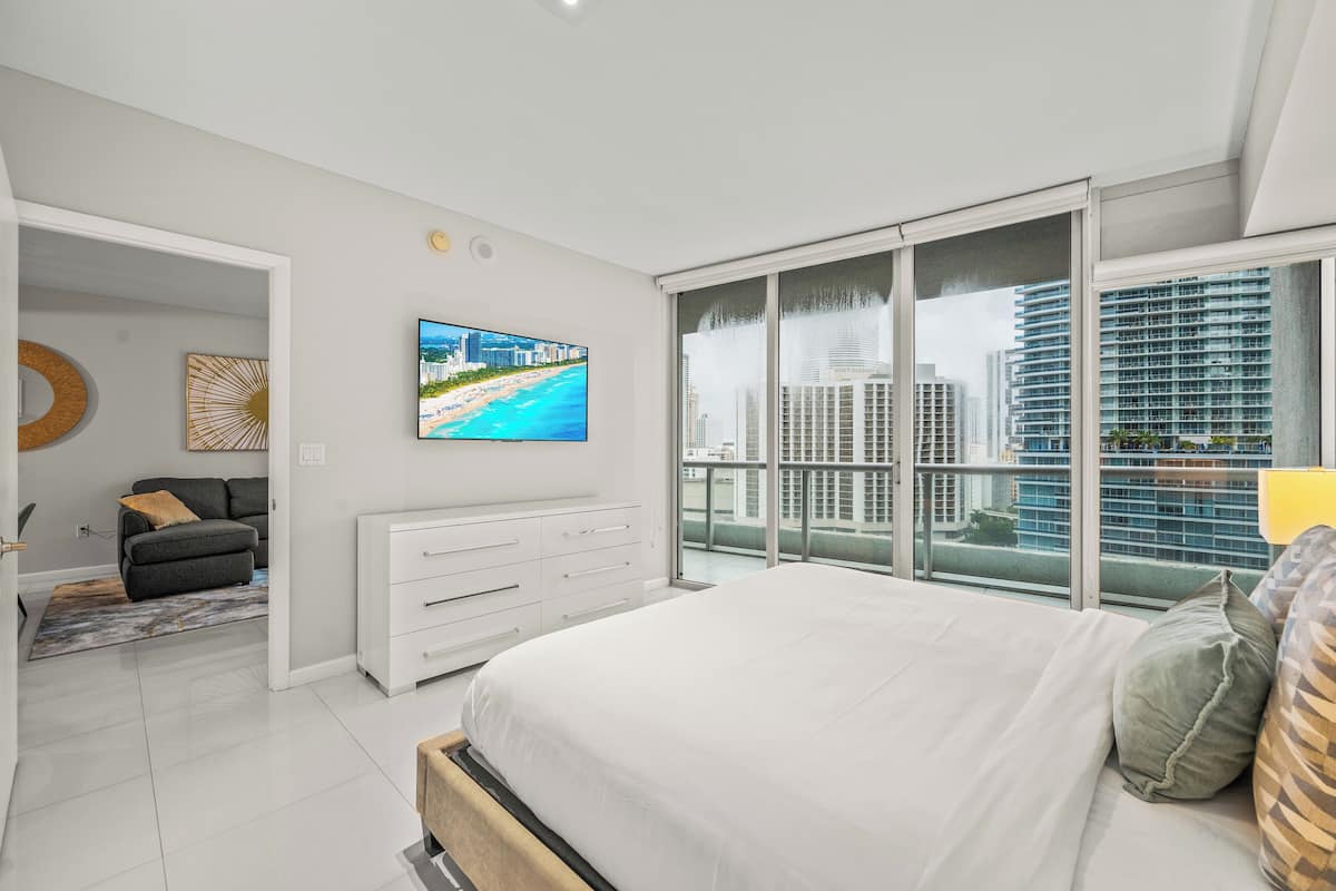 A modern bedroom with a large bed, a TV on the wall, and a balcony with city views.