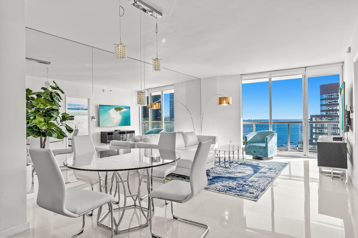 A modern living room with a glass dining table, white sofas, and a large window with a city view.