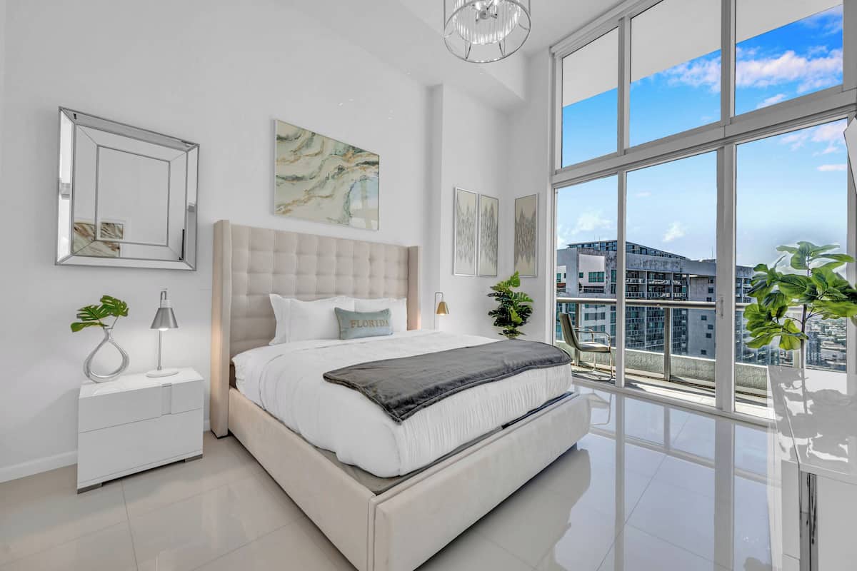 A modern bedroom with a large bed, a nightstand, a lamp, and a view of the city through large windows.
