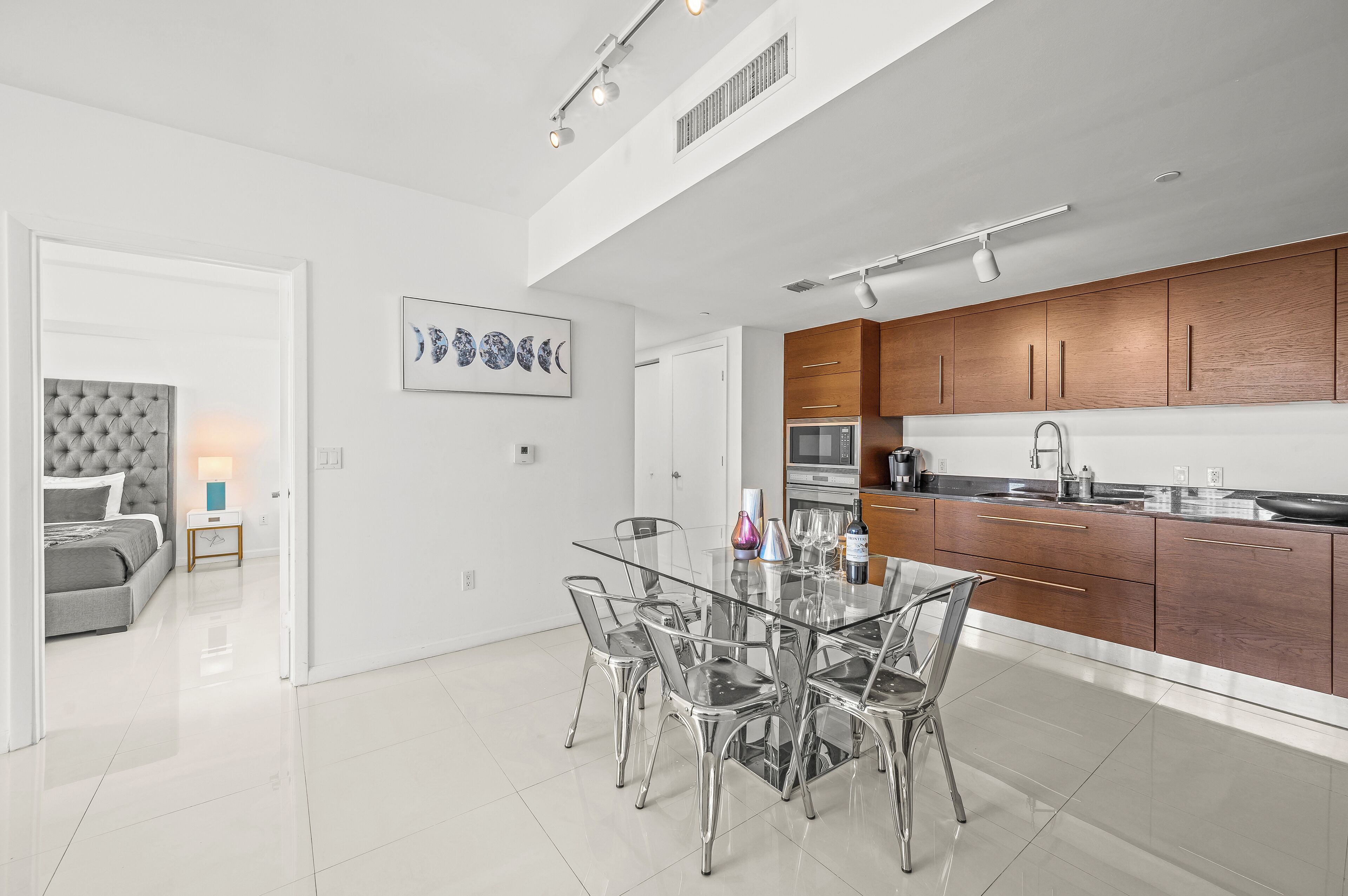 A modern kitchen with wooden cabinets, a glass dining table, and a grey upholstered sofa in the adjacent living area.