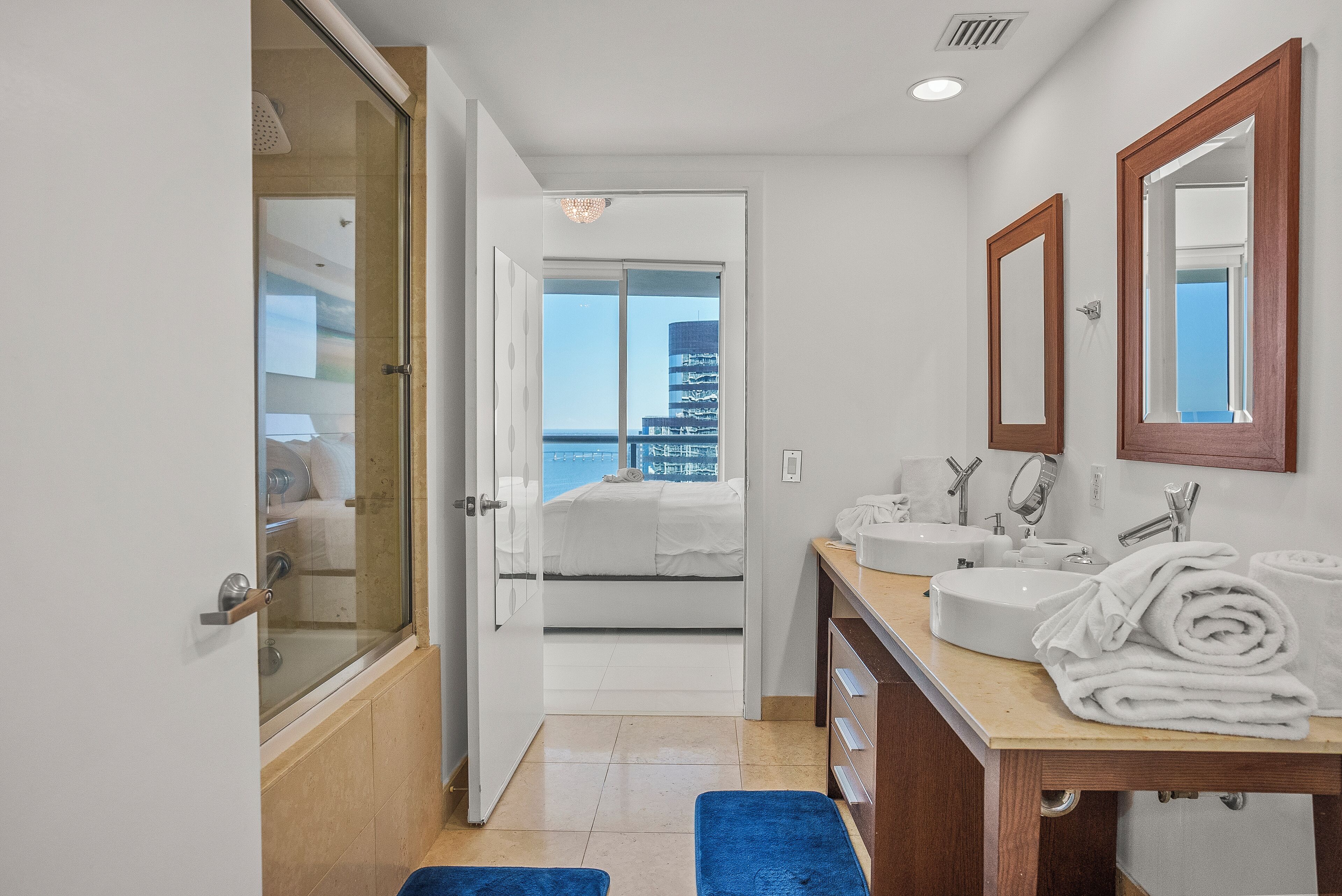 A modern bathroom with a glass shower door, a sink, a mirror, and a view of a building through the open door.