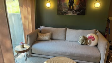 A cozy living room with a grey sofa, a small side table, and a round coffee table. There is a painting on the wall and two wall-mounted lights.