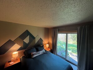 Una habitació d'hotel amb un llit gran, dues taules de nit amb làmpades, una decoració mural geomètrica i una finestra amb cortines.