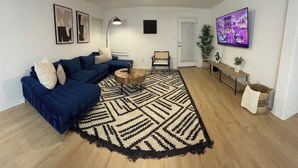 Un salon avec un canapé d’angle bleu, une table basse ronde, un tapis à motifs, un ventilateur de plafond, un téléviseur et un banc.