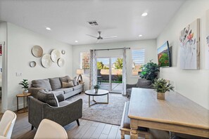 A spacious living room with a grey sofa, a wooden coffee table, and a dining table. There are decorative wall art pieces and a ceiling fan. - The Show House - Retreat for 10 (Washington)