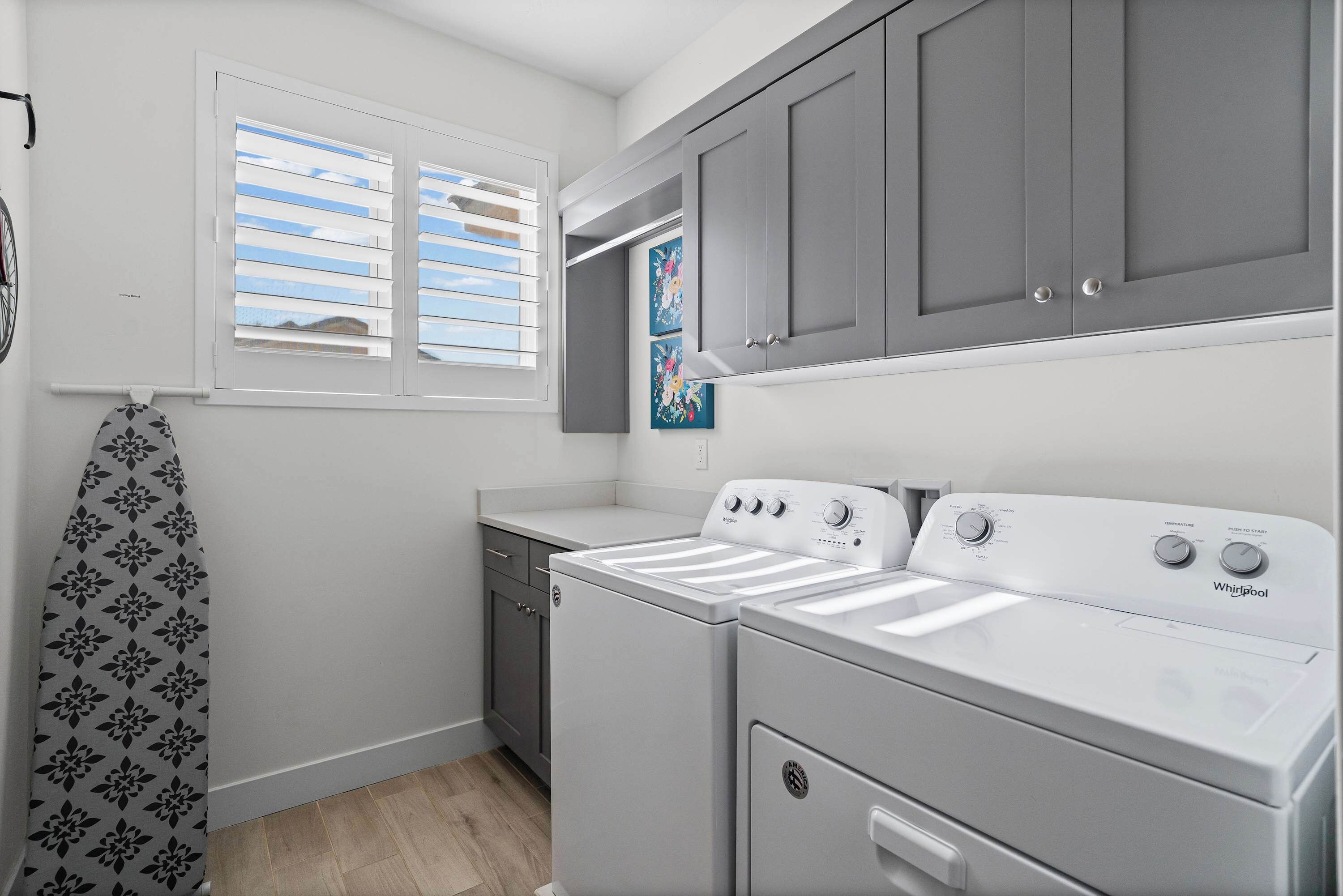 A laundry room with two white front-loading washers and dryers, grey cabinets, a window with blinds, and a patterned towel hanging on a rack.