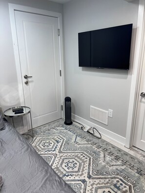 A bedroom with a bed, a small round table, a standing heater, a TV mounted on the wall, and a patterned rug.