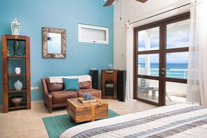 Ein Zimmer mit blauer Wand, einem braunen Ledersofa, einem Holztisch, einem Bett mit gestreifter Bettdecke, einem Holzschrank mit Dekorationen, einem Spiegel und einer großen Schiebeglasdoor mit Blick aufs Meer.