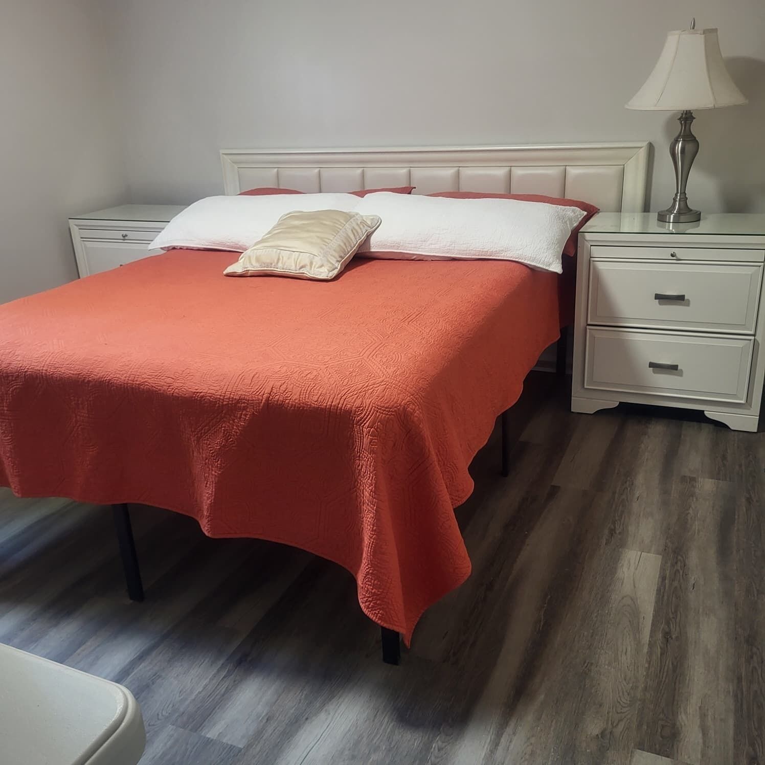 A neatly made bed with a red bedspread and two pillows, a white nightstand with drawers, a lamp, and a wooden floor.