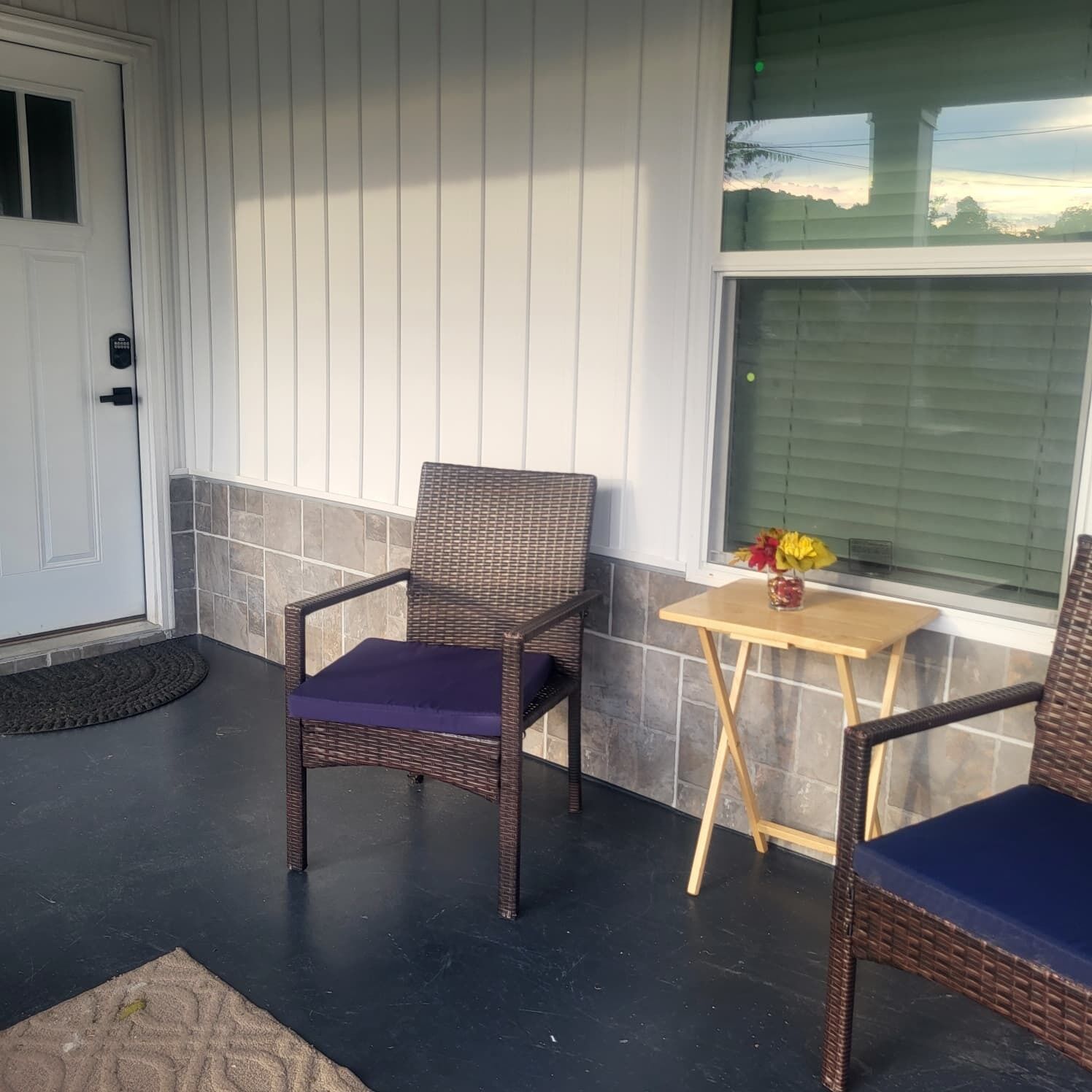 A porch with two wicker chairs, one with a purple cushion, and a small wooden table with a vase of flowers.