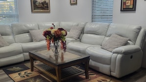 A living room with a sectional sofa, a coffee table with a vase of flowers, patterned rugs, and framed artwork on the walls.