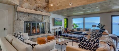 Amplia sala de estar con chimenea de piedra, techo de madera y grandes ventanales con vista a un cuerpo de agua.