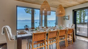 Un comedor con una mesa dispuesta para seis personas, ventanas amplias con vista al mar y dos lámparas colgantes de mimbre.