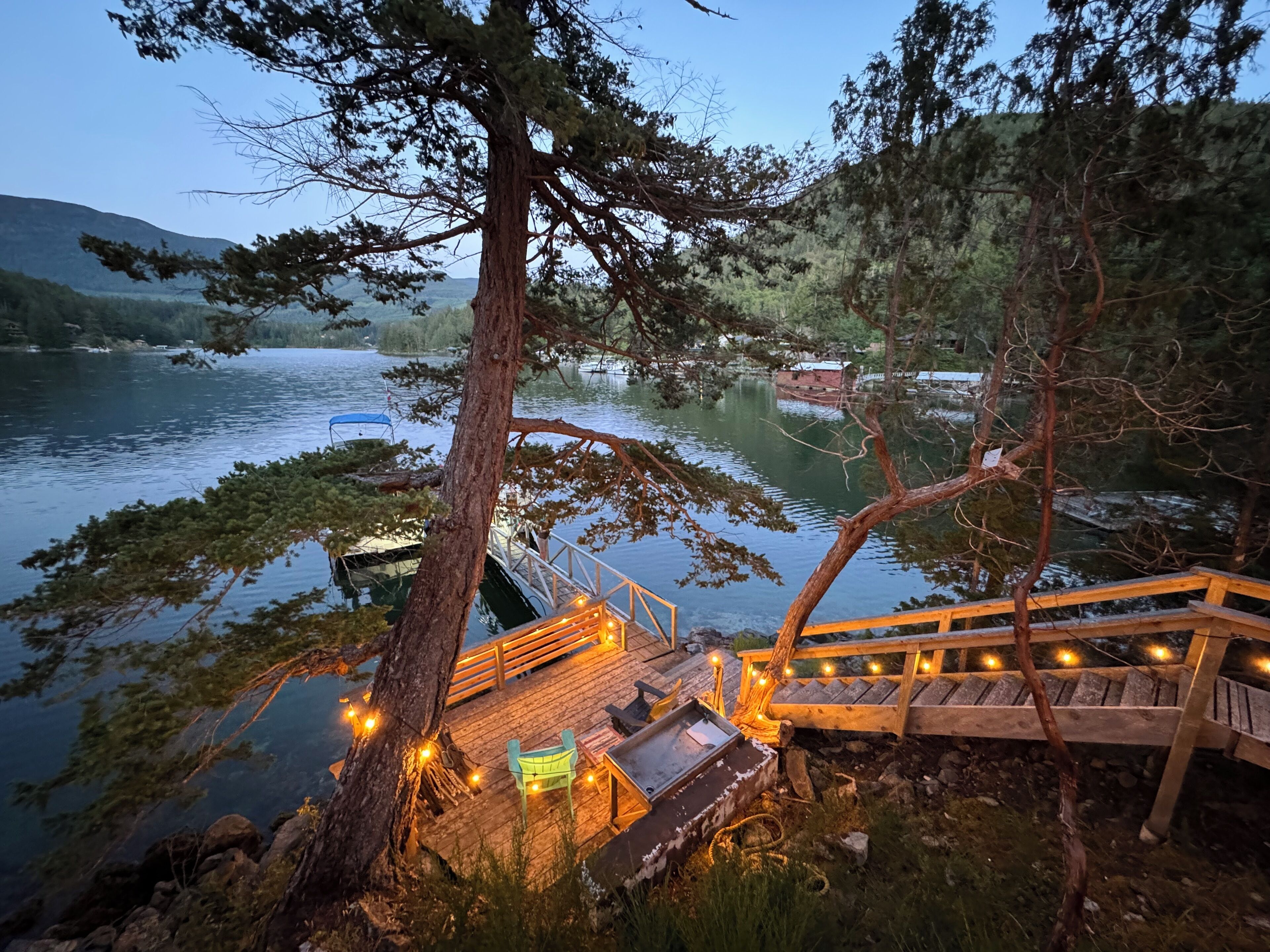 En træterrasse med gelænder, bænk og et lille bord, med udsigt over en rolig sø med både og en bro i baggrunden.