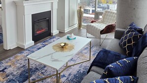 A modern living room with a fireplace, a flat-screen TV mounted above it, a sofa with blue and gold cushions, and a large window offering a city view. - Luxe 1-Bed Corner Suite in Music Row Nashville - Coworking, Gym, Skyline Views (Nashville)