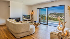 A living room with a sofa, TV, and a view of a hilltop town through large glass doors.