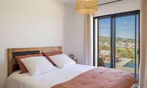 A bedroom with a large bed, a wooden headboard, a hanging light fixture, and a view of a pool and buildings through a large window. - Villa Pietra Mare, piscine vue mer, à l'Ile-Rousse (L'Ile-Rousse)