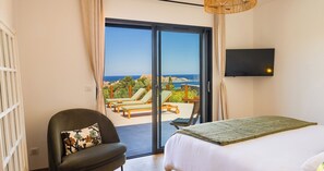 A bedroom with a bed, a chair, a TV, and a sliding glass door leading to a balcony with lounge chairs and a view of the sea.