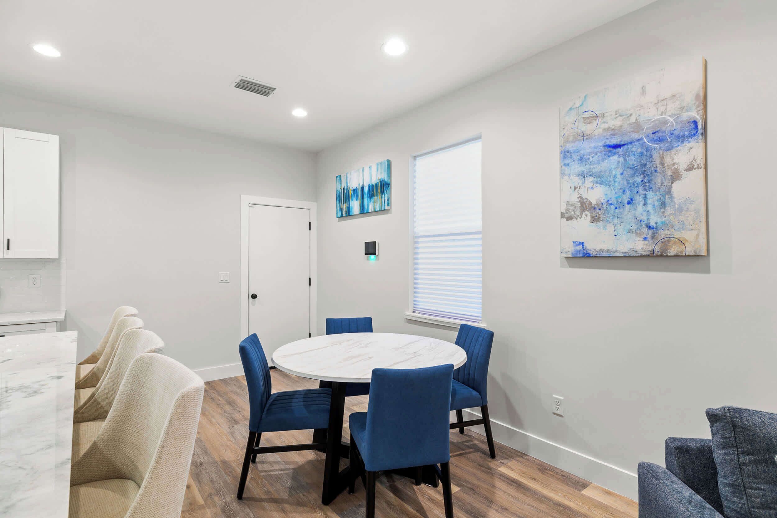 A modern dining area with a round table and blue chairs, a white door, and a large abstract painting on the wall.