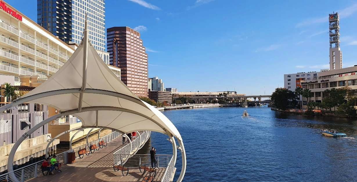 Een wandelpad langs de rivier met een overdekte zithoek, een brug op de achtergrond en een stadsbeeld met hoge gebouwen.