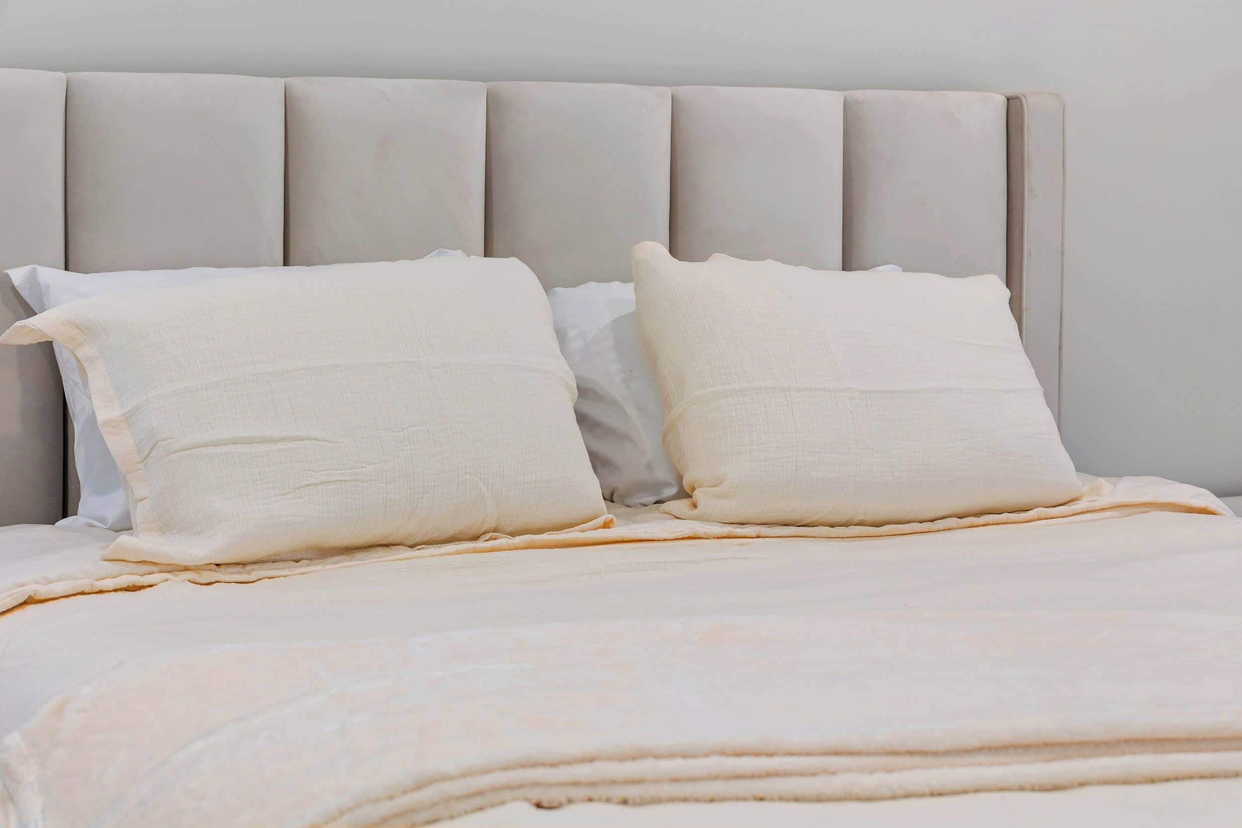Een bed met een grijs bekleden hoofdbord, twee witte kussens en een beige deken.