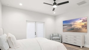 Une chambre à coucher avec un lit, un ventilateur de plafond, une commode blanche, une chaise et un grand tableau représentant une scène de plage.