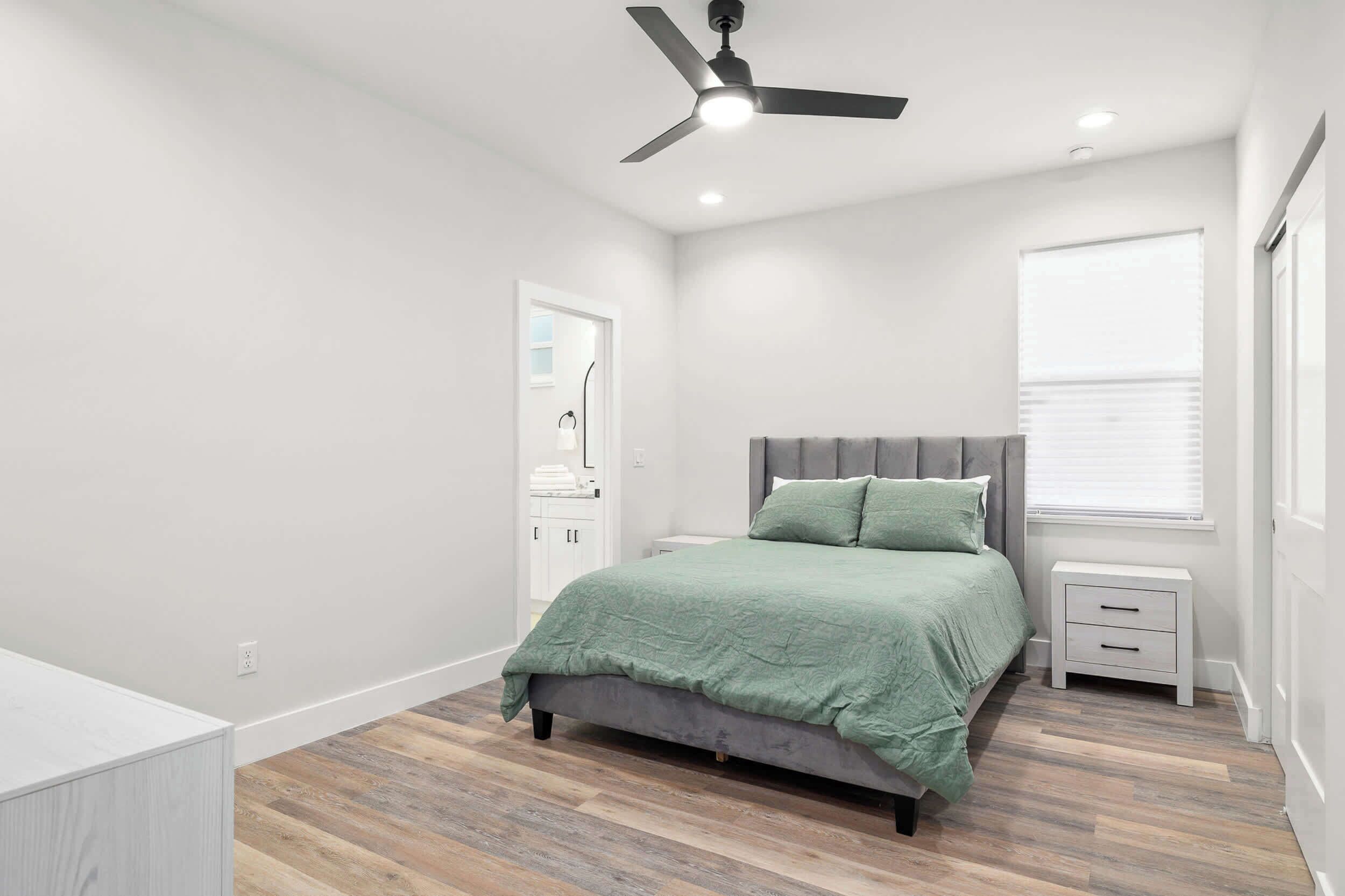 Une chambre à coucher avec un lit, une table de chevet, un ventilateur de plafond et une fenêtre avec des stores.