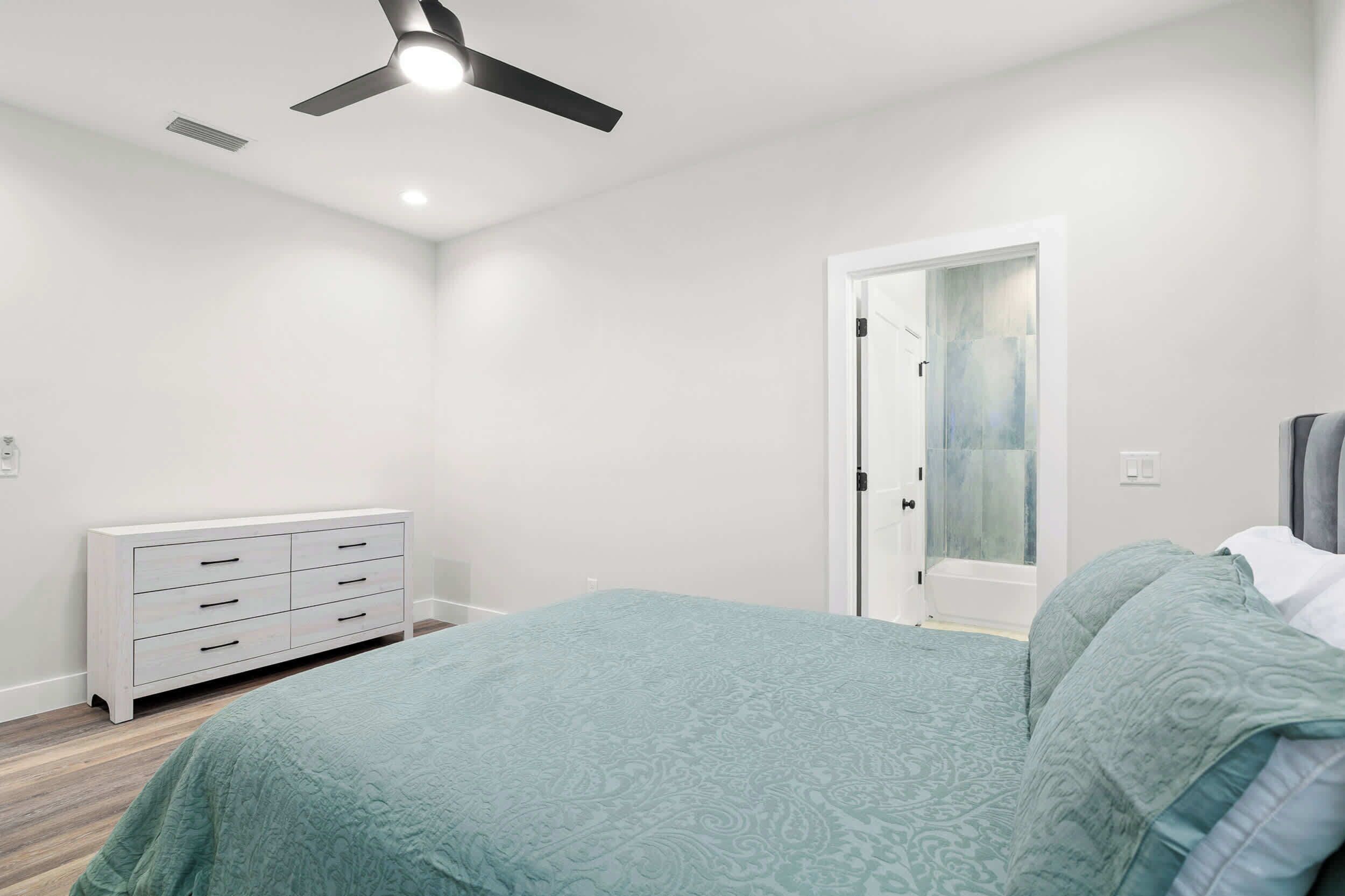 Una camera da letto con un letto, una cassettiera, una ventola a soffitto e una porta che dà su un'altra stanza.