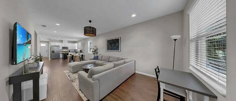 A modern living room with a grey sectional sofa, a dining table, and a TV mounted on the wall.