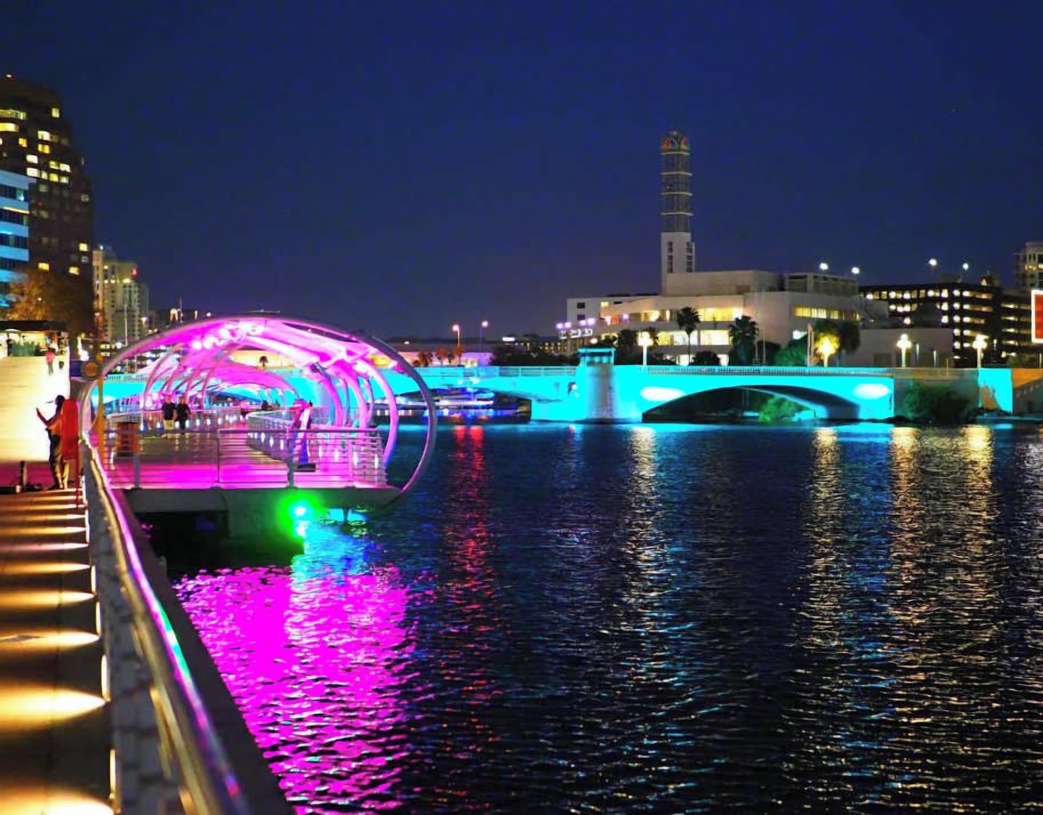 A brightly lit pedestrian bridge over water with a modern design, a tower in the background, and city buildings.