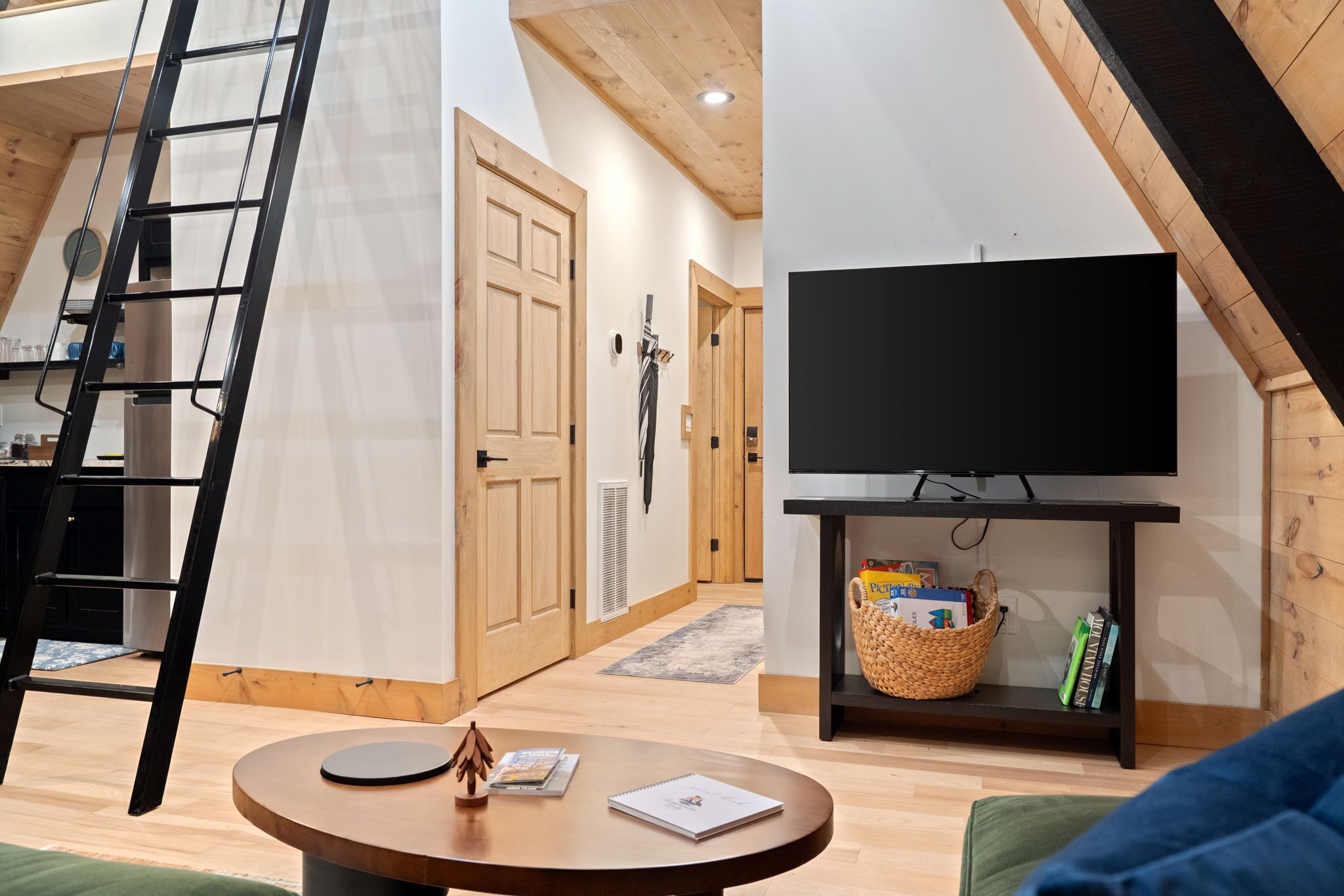 Una sala moderna con techo de madera, un televisor negro sobre un mueble bajo, una mesa redonda con libros y una pequeña figura de madera, y una cesta de mimbre en un estante.
